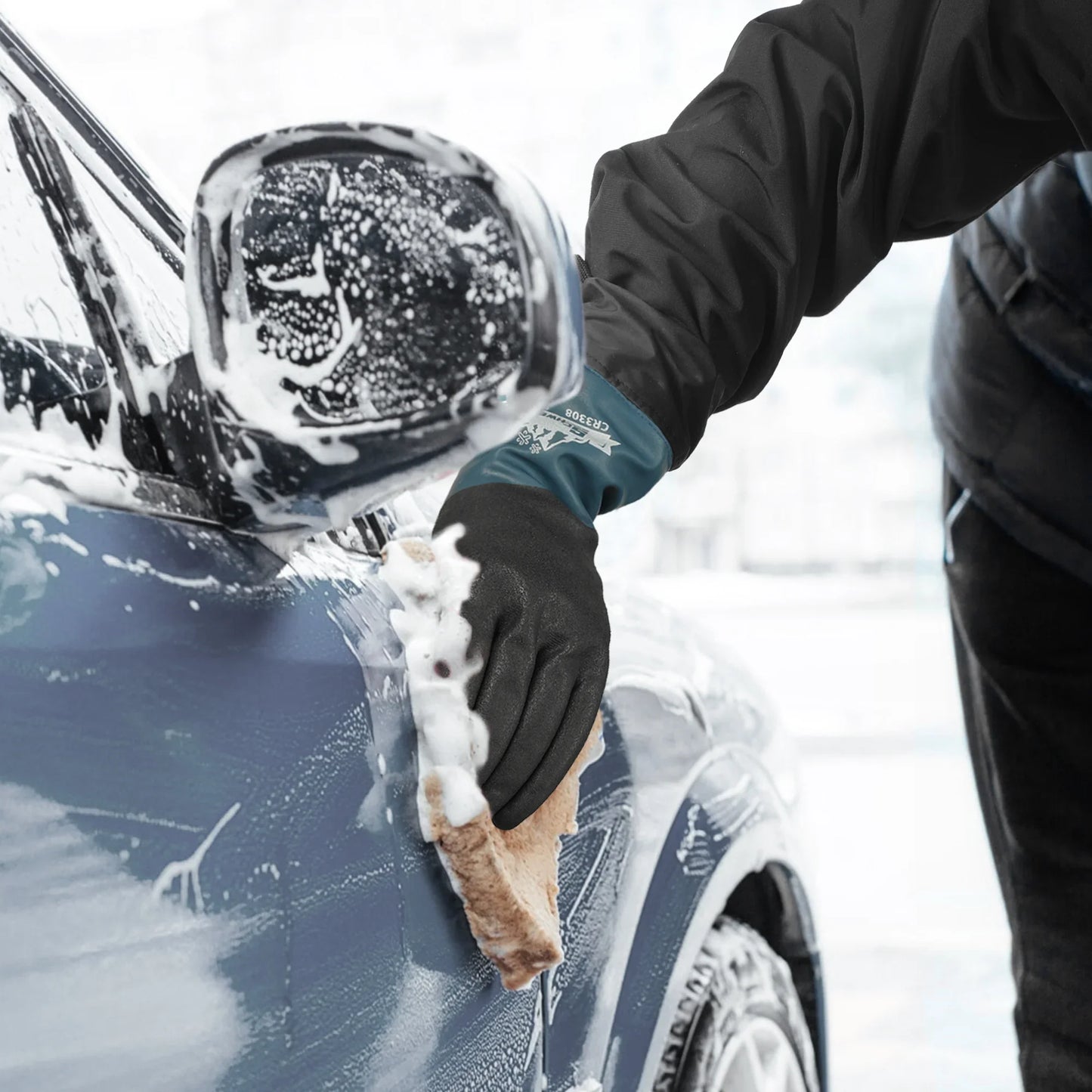 Person wearing black gloves washing blue car with sponge covered in foam near side mirror