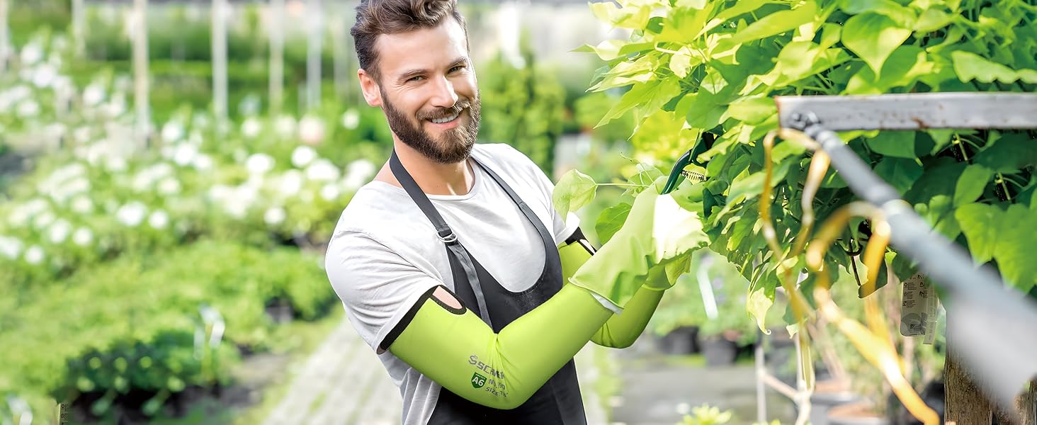 Man in a greenhouse wearing gardening gloves and an apron, tending to plants.