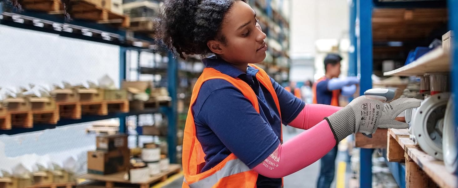 Person in a warehouse wearing an orange safety vest and gloves, interacting with equipment.