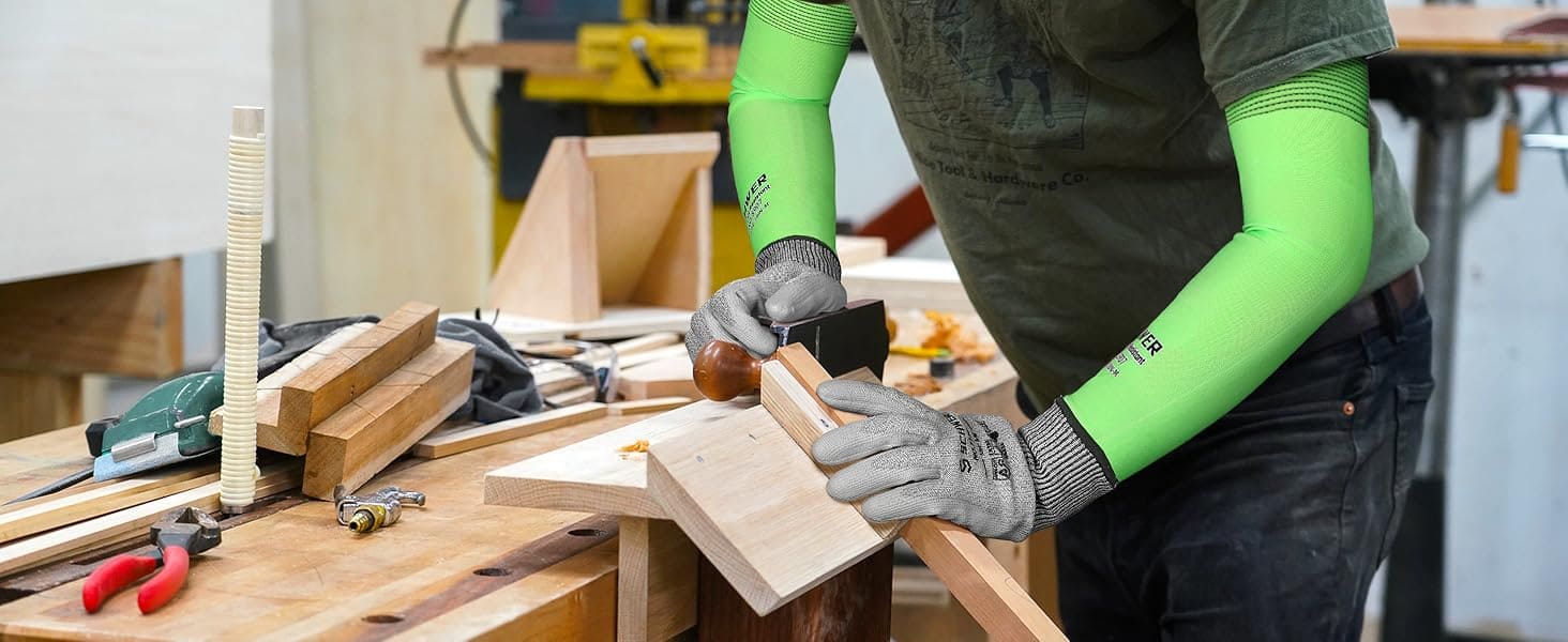 Person working with wood in a workshop wearing green sleeves and gloves.