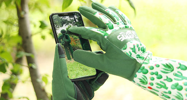 Person wearing green gardening gloves using a smartphone with a blurred natural background