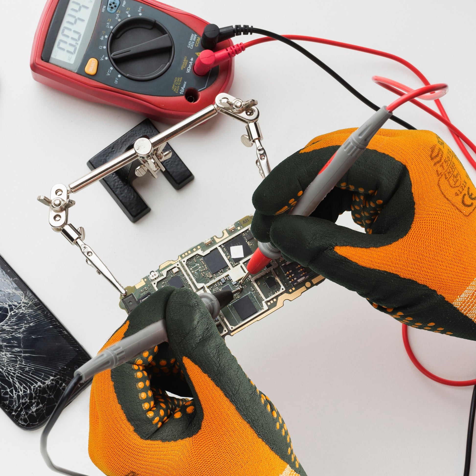 Hands wearing orange and black protective work gloves testing a smartphone circuit board with a multimeter and probes
