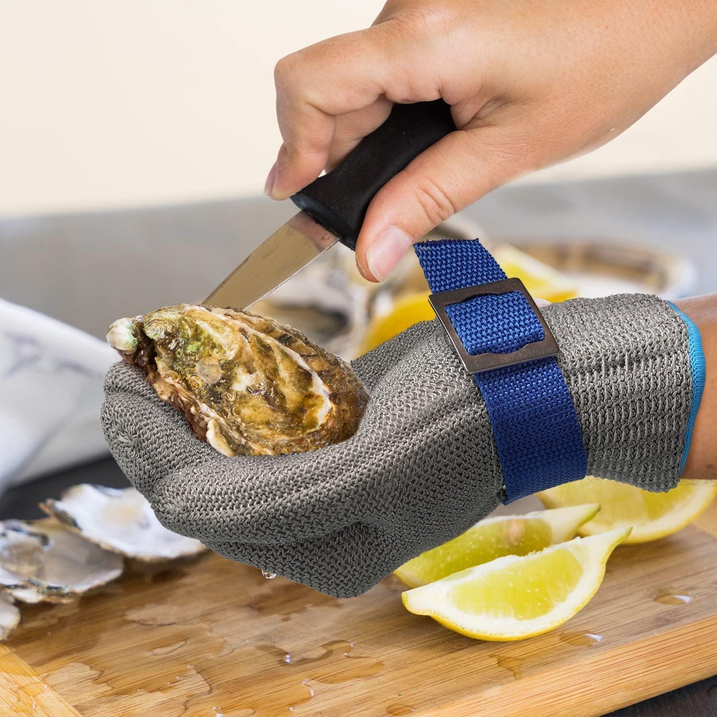 Person using an oyster shucking tool with a glove on a wooden surface with lemons.